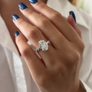 May include: A close-up of a diamond ring with an oval-cut center stone. The ring has a delicate band with smaller accent stones. The ring is set on a hand with blue nail polish, against a white shirt.