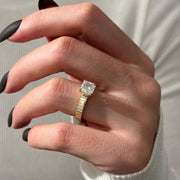 May include: A gold ring with a large, square-cut diamond. The band has a ribbed design. The ring is held up by a hand with black nail polish.